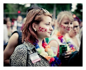 At the 2014 Gay Pride event, a young woman with short red hair and glasses perched on her head stands in the crowd, sporting "SEX" painted on her cheek. She wears a leopard print jacket and a colorful flower lei, her name tag reading "L'homophobie est circumise entourons nous." Beside her is another woman with "SEX" on her cheek.