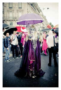 A person wearing an elaborate, purple costume with white, ornate headgear and holding a purple umbrella stands amidst a crowd on a city street. The scene is colorful with several umbrellas and people in various attires, suggesting a festive or parade atmosphere, likely during the 2014 Gay Pride celebration.
