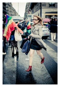 A woman in a beige trench coat and red boots hurries across a wet street during the 2014 Gay Pride parade. People behind her wave rainbow flags, and some wear colorful outfits. It appears to be a lively event in a city with tall buildings in the background.