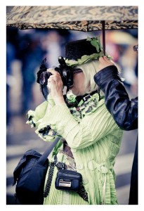 A person wearing a vibrant green, detailed outfit and a black hat is holding a camera up to their face, capturing a photo at Pride 2014. They stand under a patterned umbrella held by another individual. A black camera bag hangs from their shoulder.