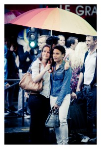 Two women stand under a large, multicolored umbrella in a crowded outdoor setting at what appears to be a 2014 Gay Pride event. The woman on the left has her arm around the shoulders of the woman on the right. Both are dressed casually, and the scene appears to be during a rainy day.
