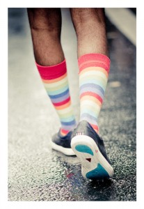 A person walking on a wet surface, wearing black shoes and colorful, striped knee-high socks that echoed the spirit of Gay Pride. The leg and foot are visible, emphasizing the vibrant socks against the muted tones of the street, reminiscent of a lively parade from 2014.