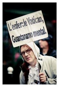 A person holding a handmade sign that reads, "L'enfer: le Vatican, Guantanamo mental," is pictured at the 2014 Gay Pride parade. The individual is wearing glasses, a white headscarf, and a light-colored jacket. The background is blurred, showcasing other participants in an outdoor setting.