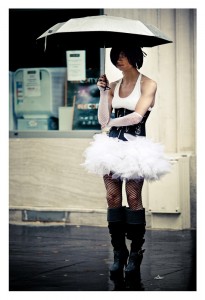 Person dressed in a white tutu, white tank top, black corset, and fishnet stockings, holding a white umbrella, standing on a wet street with a neutral expression. The background features a building window with a vending machine adorned with Pride stickers.