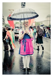 A person standing under a black-and-white umbrella wears a pink and blue dress, white tights, and black shoes. They are in a crowded outdoor area during the 2014 Gay Pride event. In the background, people carry rainbow flags, and the ground appears wet from rain.