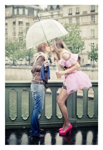 Two people are kissing under an umbrella on a rainy day. One is wearing a brown jacket and jeans; the other is dressed in a pink tutu, fishnet stockings, and bright pink heels. They stand on a bridge with an urban backdrop featuring buildings and greenery, celebrating love like it's Gay Pride 2014.