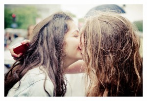 Two people with long hair share a kiss outdoors, one holding a red phone. Both appear absorbed in the moment amid a blurred backdrop, capturing the joy and love of Gay Pride.