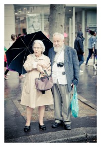 An elderly couple stands on a rainy city sidewalk during the 2014 Gay Pride parade. The woman holds a black umbrella and wears a beige coat, while the man carries a camera around his neck and a blue shopping bag. Both appear to be enjoying a moment together amidst the bustling street.
