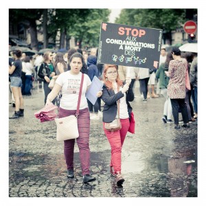 Two women march on a wet street during what appears to be a Gay Pride event in 2014, holding a sign in French that reads, "STOP AUX CONDAMNATIONS A MORT DES," followed by gender symbols. Other protestors can be seen in the background, and the participants appear determined and engaged.