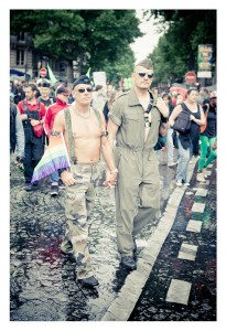 Two men walk hand-in-hand on a rainy street during a 2014 Gay Pride parade. They are wearing military-inspired outfits, one shirtless with a rainbow flag, and the other in a jumpsuit. Other parade participants are visible in the background, with trees lining the street.