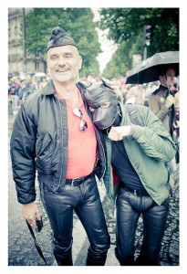 Two people stand together outdoors at a parade during Gay Pride 2014, both wearing leather outfits. One smiles, donning a black leather cap and jacket over a red shirt. The other sports a black mask and similar jacket, gesturing with a hand. Onlookers and umbrellas are visible in the background.