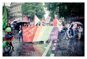 A diverse group of people march on a rainy street for Gay Pride 2014, holding a large rainbow banner that reads "L'égalité maintenant! L'égalité c'est classe" and other signs promoting equality and LGBTQ+ rights. Some participants carry umbrellas and flags.