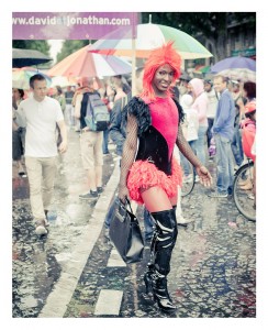 A person in a vibrant, feathered red outfit and black thigh-high boots walks through a crowded street during the 2014 Gay Pride parade. They carry a black bag and smile, surrounded by people with colorful umbrellas. A website banner is visible in the background.