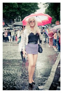 A person with long, blonde hair stands in a rainy street under a red and white umbrella during the 2014 Gay Pride. They are wearing a silver jacket, black top, and striped black and white skirt. In the background, others with umbrellas walk by alongside a vibrant pride flag.