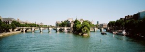 A wide view of the Seine River in Paris with the iconic Pont Neuf bridge spanning the water, lined with trees and historic buildings on both banks under a clear blue sky—a perfect scene for an été stroll.