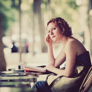 A woman with curly hair sits at an outdoor café table, her yeux thoughtful as she holds a book. A cup of coffee and a bag rest on the table. Through the blurred fenêtre behind her, a tree-lined street can be seen.