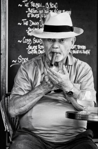 An older man in a hat, seated at a café table, lights a cigar. He wears a collared shirt and watch, with a drink nearby. Behind him, a chalkboard menu is partially visible. The image is in black and white.
