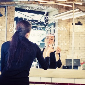 A woman with long dark hair writes on a mirror with a white marker in a tiled room. The words "NO SMOKING" are at the top, and her reflection reveals her savoir faire as she concentrates on each word.