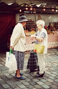 Two elderly women enjoy a conversation at the fin de marché. One carries a shopping bag and wears a hat, while the other holds a yellow handbag beside her plaid wheeled cart. Fruit stalls fill the lively background.