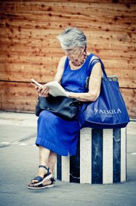 La dame en bleu, an elderly woman with gray hair, sits on a striped stone pedestal in her blue dress and sandals, reading a book. A black purse and large blue shopping bag rest at her side against the wooden wall background.
