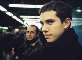 Two men stand on a crowded subway platform. One, in the foreground, gazes neutrally at the camera; the other faces forward. Both wear dark clothing and scarves. The scene is rendered in warm Kodak Portra tones.