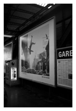 A black and white photo captures an indoor public area with a large poster on the wall. The poster depicts a person falling or jumping with buildings in the background. Nearby, there's a vending machine and an information board labeled "GARE" (station). Below, "Octobre 2014" suggests a contemporary moment akin to Garry Winogrand's visual narratives.