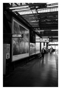 A black and white photograph of an RATP train station platform with large advertisement posters on the wall, captured in October 2014. A sign with "Alarme" is visible. One person wearing a suit and carrying a bag is walking along the platform. The station has a modern roof structure reminiscent of Garry Winogrand's style.