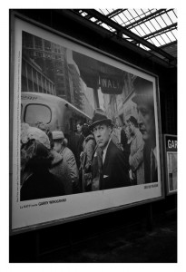 A black-and-white photograph displayed on an RATP billboard shows a crowded urban scene with people walking. In the center, a man in a suit and hat looks directly at the camera. There is a bus and buildings in the background. The text on the billboard includes "GARRY WINOGRAND, Octobre 2014.