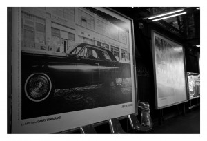Black and white photograph of a subway station with large posters on the wall. One prominent poster features a classic car and the name Garry Winogrand, alongside an ad for Octobre 2014 by RATP. The station has empty plastic chairs and dim lighting, creating a nostalgic and moody atmosphere.