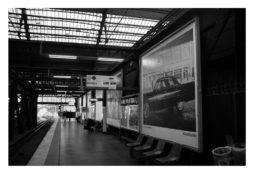 A black-and-white photo of a train station platform with a high glass ceiling. There are several posters along the walls, including a large one of a classic car. A few people are waiting on benches, and the track extends into the distance on the left, evoking scenes reminiscent of Garry Winogrand’s candid moments.