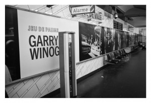 A black and white photograph of a subway station with a long wall displaying a large sign that reads "Jeu de Paume Garry Winog." The wall, adorned with black and white images taken by Garry Winogrand, has empty chairs lined up beneath. A sign above reads "Alarme," marking the RATP station in Octobre 2014.