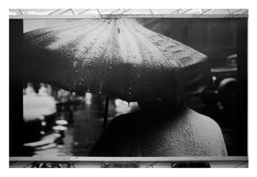 Black and white photo depicting a person holding a large umbrella with raindrops on it, walking on a wet street. The person's back is visible, and they wear a coat. The background, reminiscent of Garry Winogrand's style, shows blurred shadows of buildings and RATP vehicles, enhancing the rainy atmosphere.
