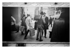 A black and white photograph reminiscent of Garry Winogrand's work captures a busy city street scene from the mid-20th century. Several men, dressed in suits and hats, stand and walk along the sidewalk. One man carries a briefcase while another carries a box. Buildings and storefronts line the street.