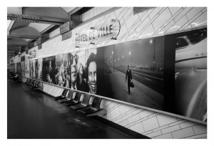 A black-and-white photograph of the Hôtel de Ville subway station. The station walls, managed by RATP, are adorned with large, vintage photos reminiscent of Garry Winogrand’s style, featuring close-ups of smiling faces, a man running with a briefcase, streetlights, and an old car. The station is empty with metallic seats lining the wall.