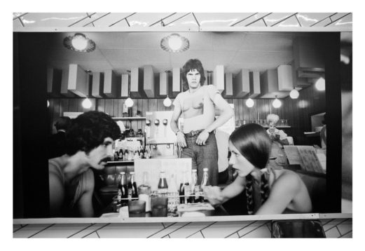 A black-and-white photo by Garry Winogrand shows a man standing shirtless at a diner counter, looking forward with a serious expression. In the foreground, a man and woman sit at a table with drinks and food, appearing engaged in conversation. The diner has vintage decor reminiscent of RATP stations.