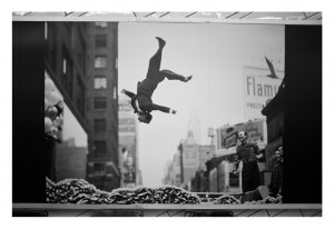 A black-and-white photo by Garry Winogrand shows a man in mid-air performing a backflip over a large pile of leaves on a city street. Two observers in uniformed RATP attire stand to the side, watching. Tall buildings and a billboard are visible in the background.