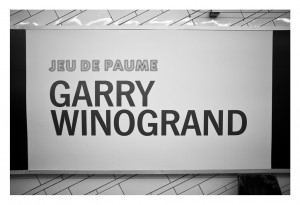 A large white wall has text that reads "JEU DE PAUME" in dotted font and "GARRY WINOGRAND" in bold black letters. The wall is part of an exhibit space with a minimalist design featuring diagonal lines, showcasing the RATP collaboration for the Garry Winogrand exhibit from Octobre 2014.