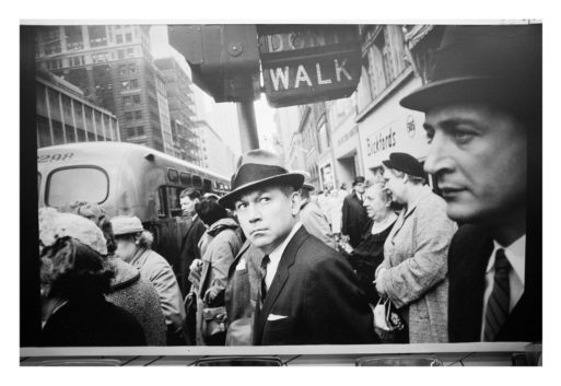 A black-and-white photo of a bustling city street reminiscent of Garry Winogrand's style. Several people, mostly wearing hats and formal clothing, wait at a street corner near an "RATP" sign. A bus is visible in the background. The atmosphere appears mid-twentieth century, with a notable man in a suit and hat glancing toward the camera.