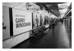 Black and white photo of a subway station with a man walking along the platform. The wall is adorned with a large advertisement for an Octobre 2014 "Garry Winogrand" exhibition, featuring a series of black and white photographs. The station is "Hôtel de Ville," maintained by RATP.