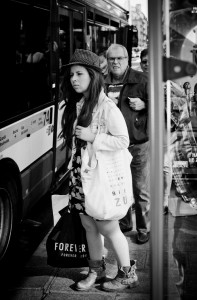 A young woman with long hair and a hat stands at an arrêt du bus, carrying shopping bags. People wait behind her to board. The scene is in black and white, set on a bustling city street with the bus in the background.