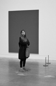 A woman stands, tourner le dos to a large, solid-colored art moderne painting in a gallery. She wears a coat, sneakers, and carries a patterned bag. The scene is in black and white.