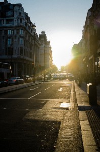 A sunlit Oxford Street city scene with cars and buses in traffic, tall buildings on both sides, and sunlight streaming in from the horizon, casting long shadows on the pavement.