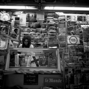 A person with curly hair sits behind the counter of a newsstand, surrounded by neatly stacked newspapers and magazines in French, displayed on shelves and racks. The scene is in black and white.