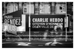 A black and white photo of a city street shows large signs reading "PARIS RENDEZ-VOUS" and "CHARLIE HEBDO, CITOYEN D'HONNEUR DE LA VILLE DE PARIS" in front of a historic building.