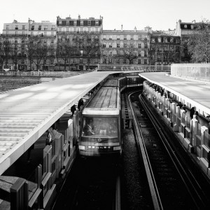 Black and white photo of a metro train arriving at Métro Bastille, with people waiting on both platforms and classic Parisian apartment buildings in the background.
