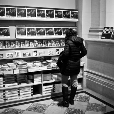 A person in a winter coat browses books and postcards on display in a Paris Magnum bookstore or museum gift shop. The scene is in black and white.