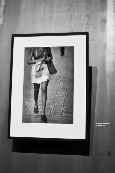 Black-and-white photo of a framed picture on a wall, showing the lower half of a woman walking in Paris, holding a shopping bag. The artist’s name and Magnum details are printed on the wall beside the frame.
