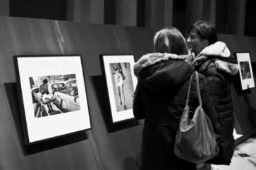Two people in winter coats stand closely together, viewing framed black-and-white Magnum photographs displayed on a gallery wall. One person is smiling. The Parisian scene is captured in striking black and white.
