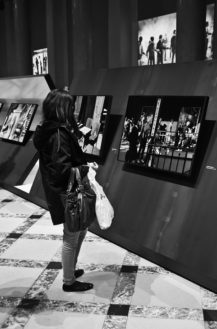 A woman stands in a Paris gallery, holding a handbag and a plastic bag, viewing framed black-and-white photographs by Magnum on angled panels. The patterned floor adds texture to the striking black-and-white scene.