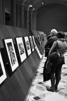 People view framed photographs displayed on angled black panels in an art gallery. The marble floor, tall columns, and archways evoke a Paris ambiance—an elegant setting for this Magnum-inspired black and white image.